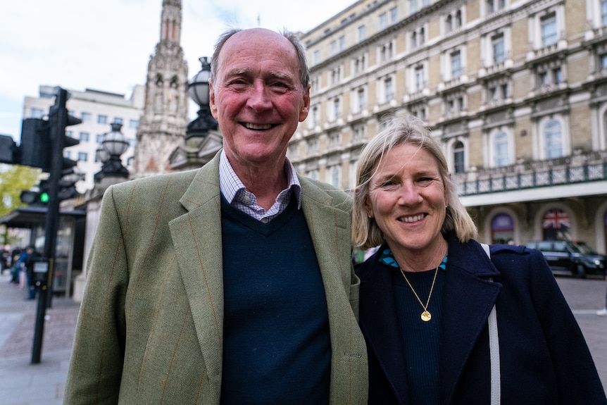 A tall man in a green jacket and a blonde woman in a black jumper smile at the camera on a London street.