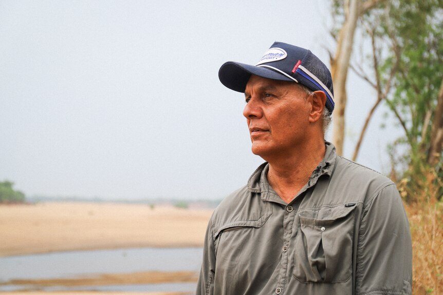 A man wearing a cap and grey shirt, standing outside near sand and water.