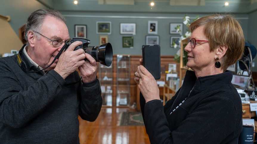 a man holds an analogue camera and a woman holds an iphone they are pointing them at each other