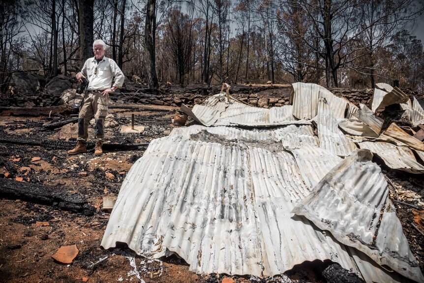 Man with camera stands beside flattened house and burnt trees