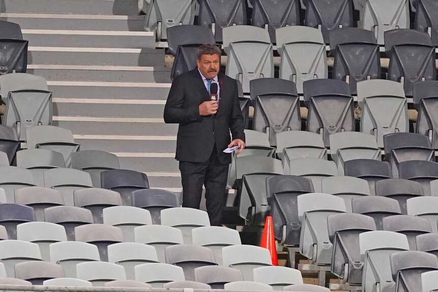 A TV commentator stands in an empty grandstand holding a microphone during an AFL match.