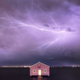 Bolt of lighting across purple-coloured stormy sky out at sea over a shed in the centre foreground.