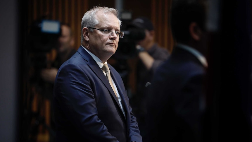 Morrison seen looking to right of frame, hands clasped at front. Out-of-focus cameras in background.