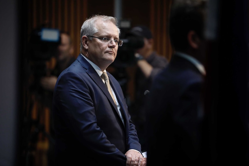 Morrison seen looking to right of frame, hands clasped at front. Out-of-focus cameras in background.