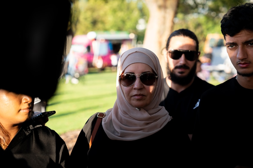 A woman in a headscarf and glasses stands in a park looking at the camera.