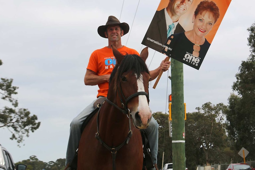 A man wearing an orange One Nation t-shirt rides a horse holding a sign with a photo of his face and Pauline Hanson.