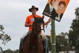 A man wearing an orange One Nation t-shirt rides a horse holding a sign with a photo of his face and Pauline Hanson.