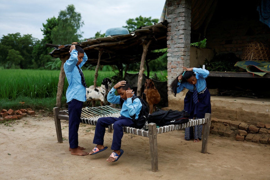 Two boys and a woman stand outside a brick hut in blue school uniforms, all brushing their hair. Green fields and forest behind