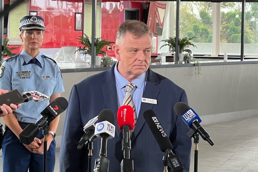 A man stands in front of microphones, with a policeman behind him 