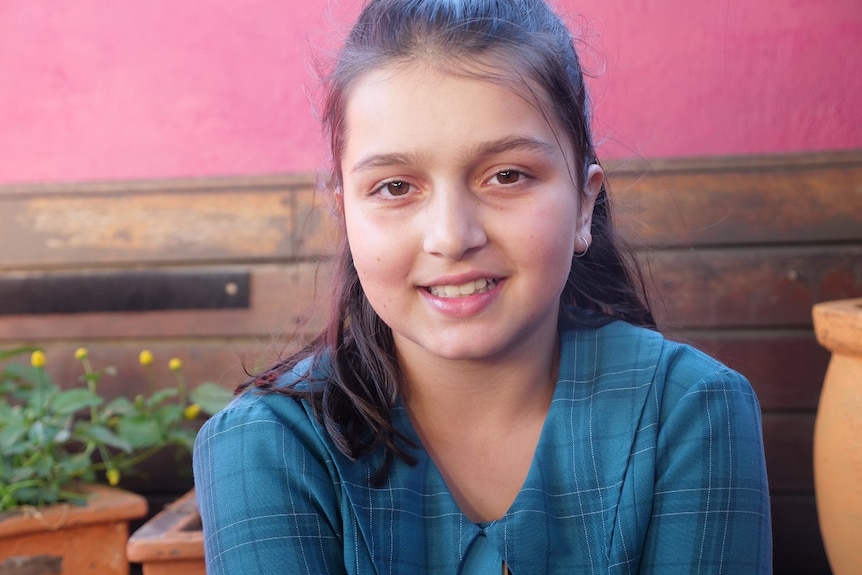 A close up shot of Priya smiling, flowers and a wall behind her.