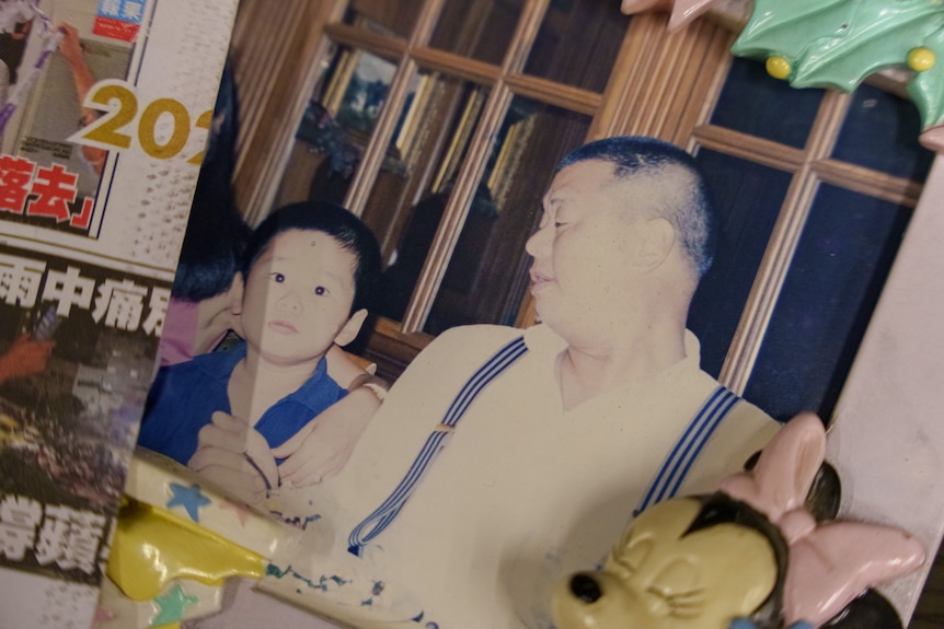 A little boy with black hair sits next to his father wearing a white shirt and suspenders.