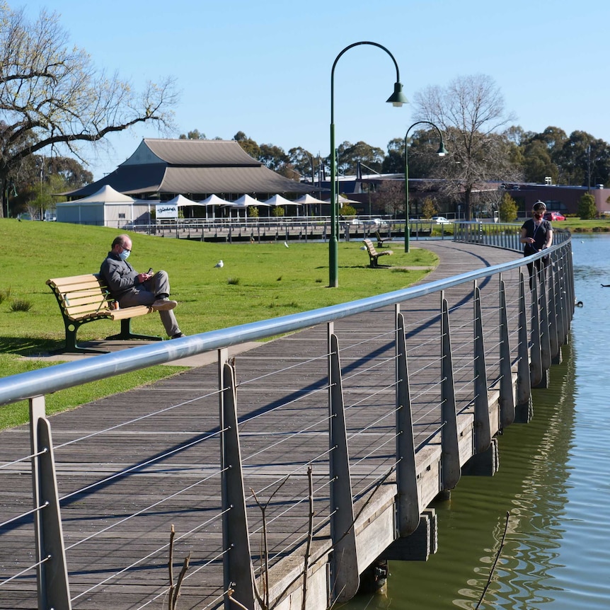 A man wearing a mask sits on a park bench beside a path opposite from a lake.