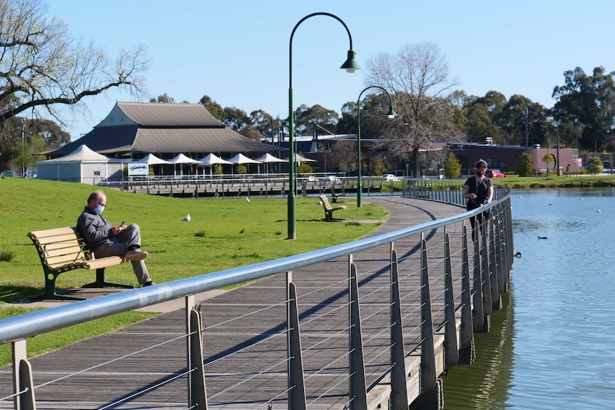 A man wearing a mask sits on a park bench beside a path opposite from a lake.