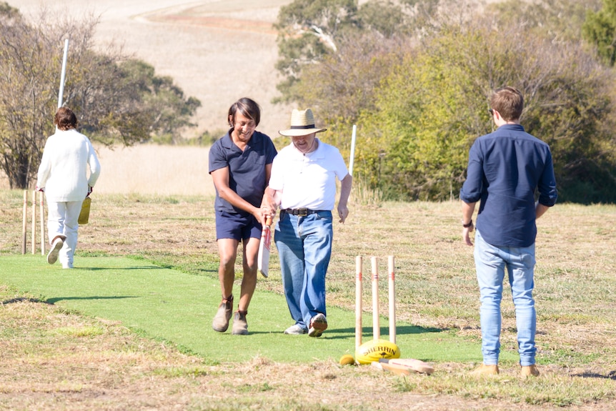 A man holding a cricket bat on a pitch with another man by his side helping him navigate