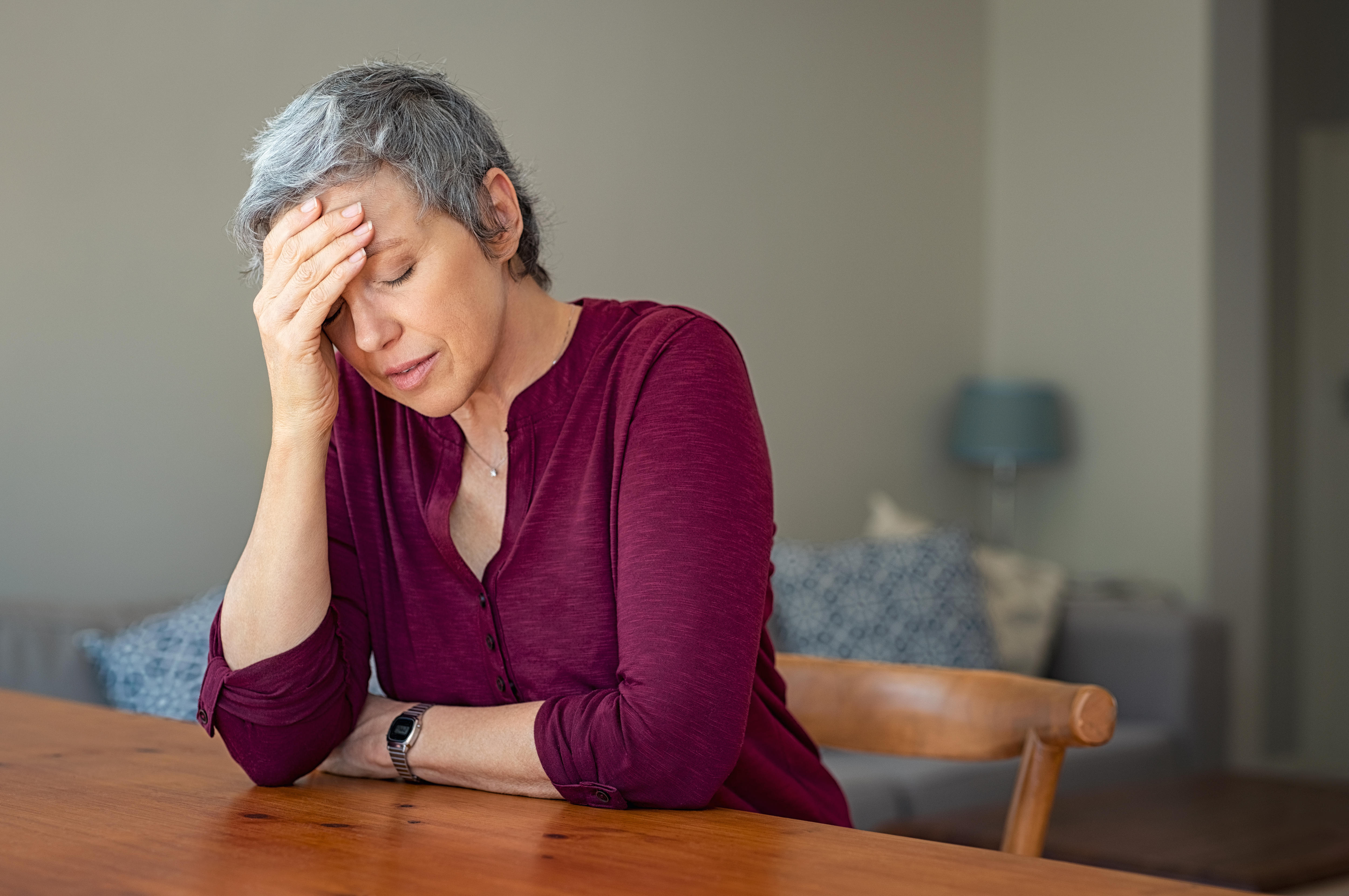 A stressed-looking woman with short gray hair and maroon top sitting at a table with her head in her hand.