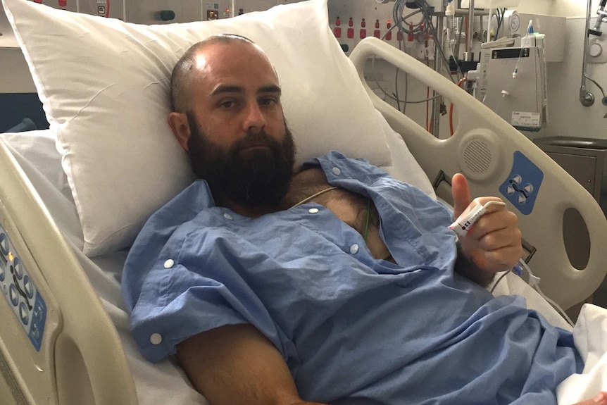 A man in blue medical gown giving a thumbs up  in a  hospital bed.