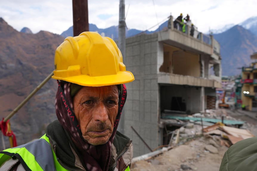 A man wearing a yellow helmet looks towards the camera. A building can be seen in the background. 