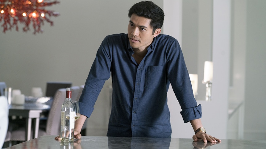 Colour still of Henry Golding in blue shirt leaning on marble table top inside a house in 2018 film A Simple Favour.