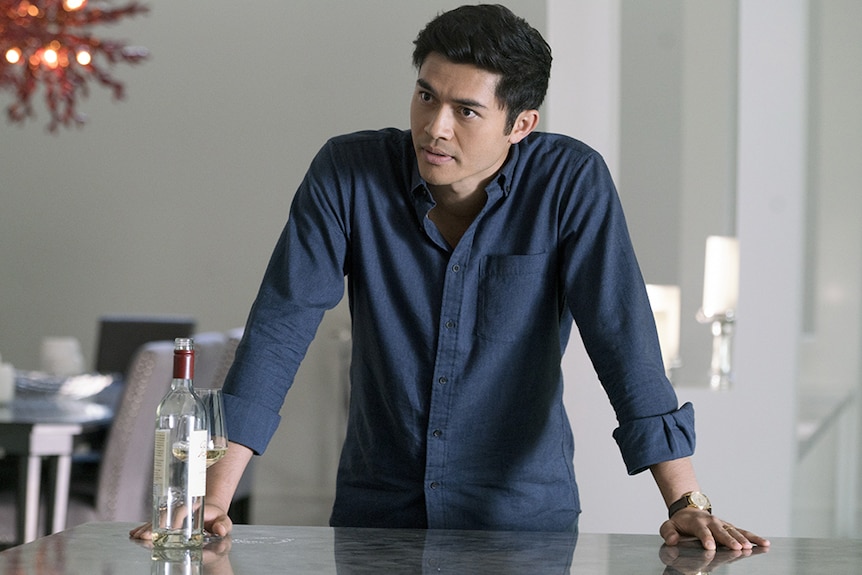 Colour still of Henry Golding in blue shirt leaning on marble table top inside a house in 2018 film A Simple Favour.