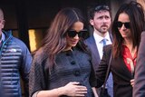 Meghan wears a black coat and sunglasses and looks down as she holds her baby bump with a grey bag on her arm.