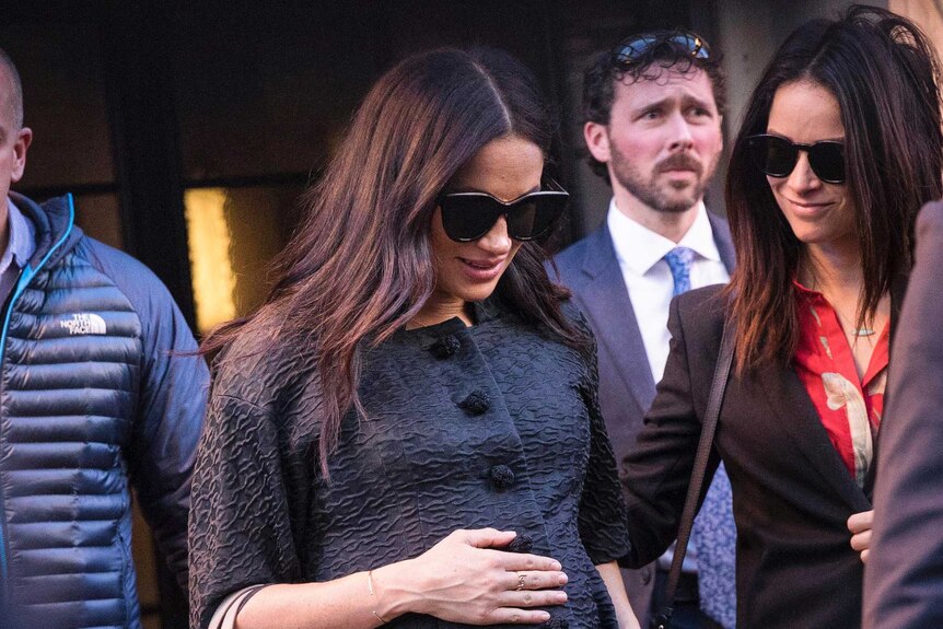 Meghan wears a black coat and sunglasses and looks down as she holds her baby bump with a grey bag on her arm.