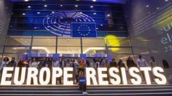 A sign reads "Europe Resists" with a blue light above. People stand behind it on steps.