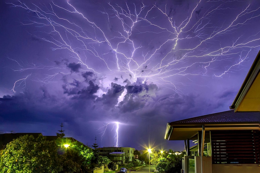 A single bolt of lightning strikes the ground as numerous bolts arc across the night sky, illuminating clouds.