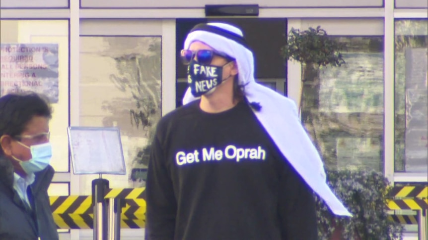 A man wearing a face mask which says 'fake news' and a t-shirt which says 'Get Me Oprah'.