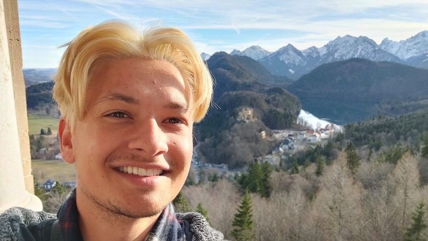 Luke Marin standing on a balcony taking a selfie in front of an alpine valley with snow cover peaks in the distance.