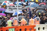 A packed crowd of protesters in wet weather gear and umbrellas stand face-to-face with orange jacketed police