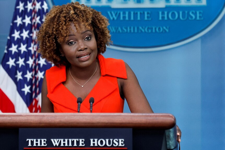 A middle-aged woman with curly hair wearing a red top standing at a podium that says the white house