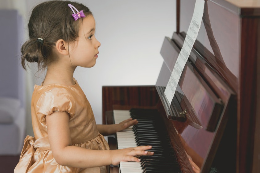 A photo of a small child sitting at an upright piano looking at sheet music.