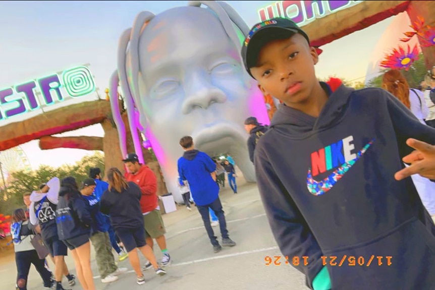 A young African American boy shows two fingers as he poses in front of a theme park with people behind