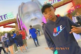 A young African American boy shows two fingers as he poses in front of a theme park with people behind