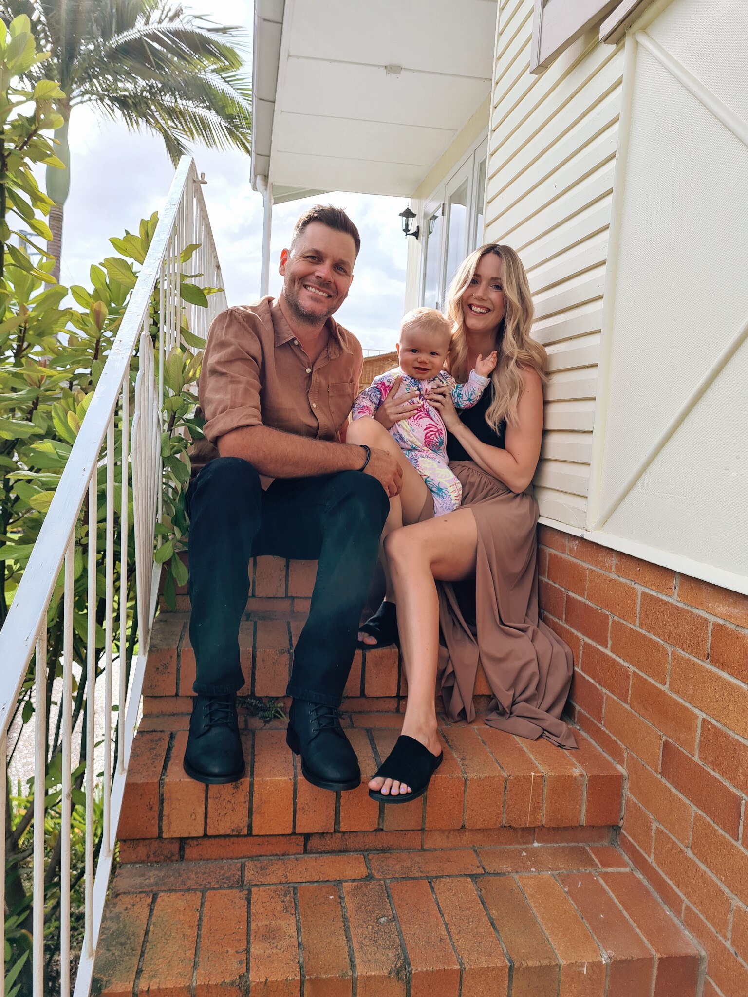Travis, Kellie and baby Lane sit on the steps of a house.
