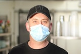 A man stands in a commercial kitchen with sunlight on his facing smiling and wearing a face mask. 