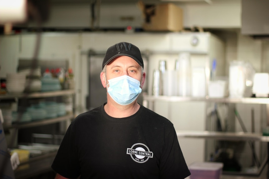 A man stands in a commercial kitchen with sunlight on his facing smiling and wearing a face mask. 