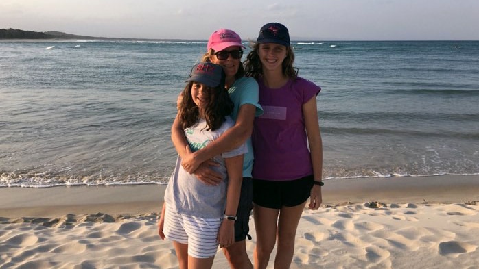 A woman hugs the girl in front of her, while another girls stands by her side at the beach.