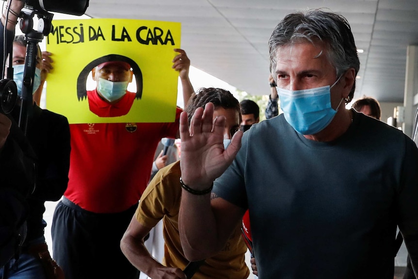 A man wearing a mask holds his hand up as he stands in front of a camera in an airport.