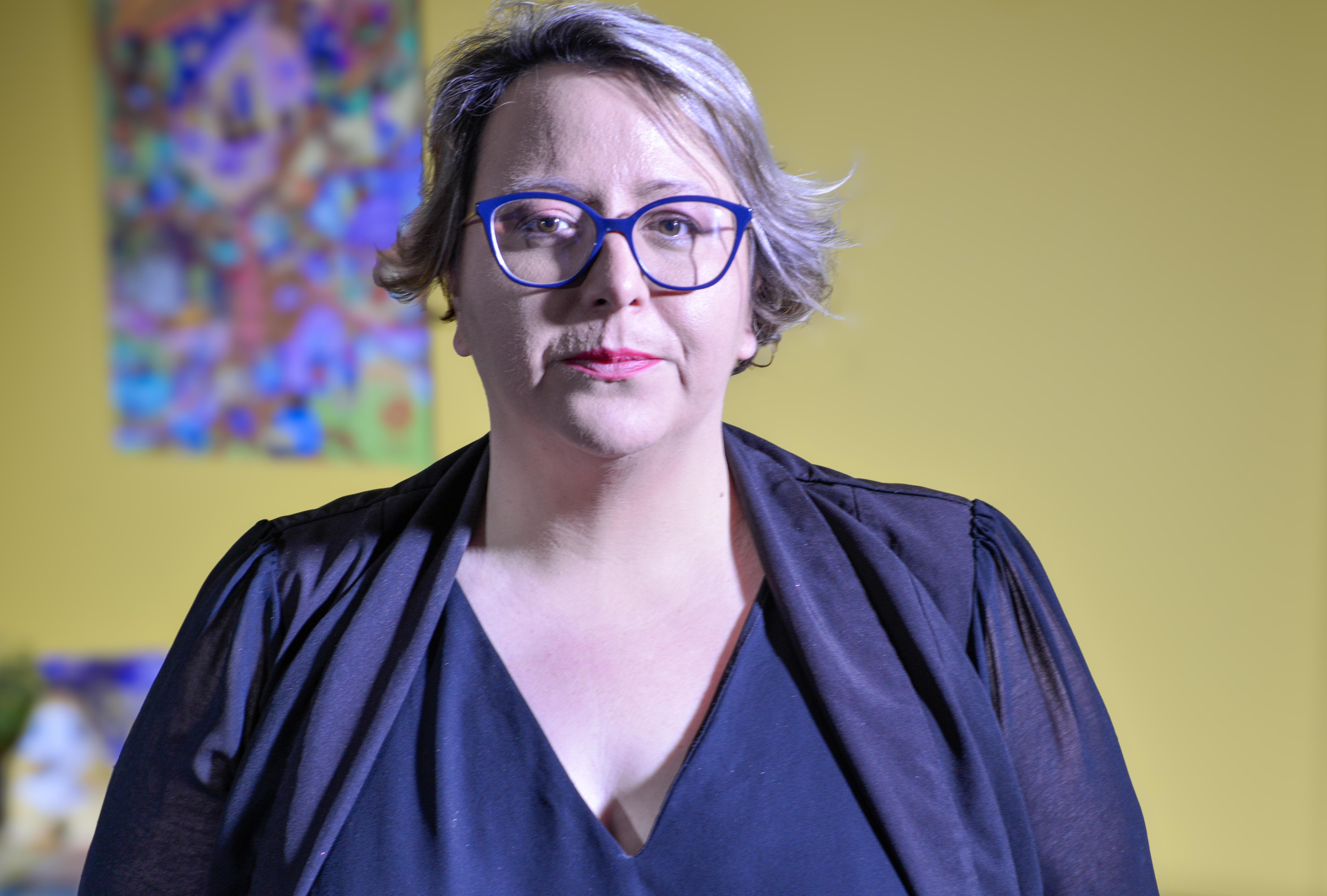 A woman with glasses looking serious in front of a yellow wall