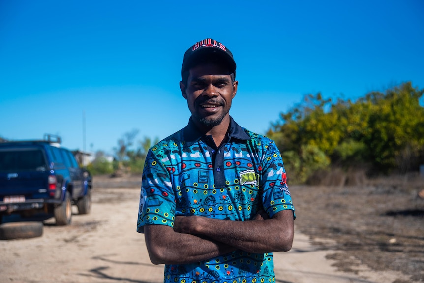 A young man wearing a hat and a blue shirt stands with his arms crossed with the blue sky and trees in the background. 