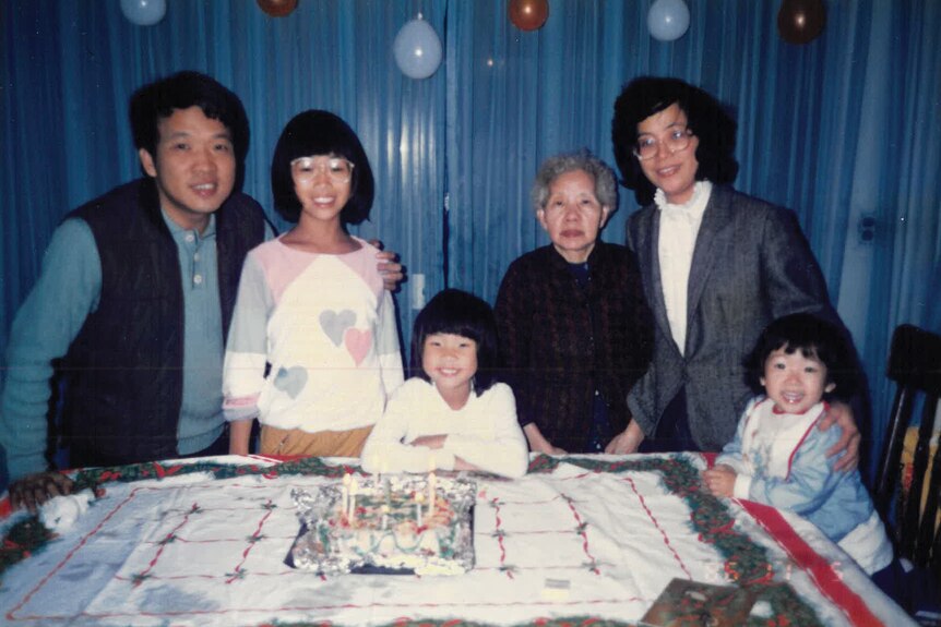 Slightly grainy family photo of young girl smiling in front of large cake, with siblings, parents and grandmother surrounding.