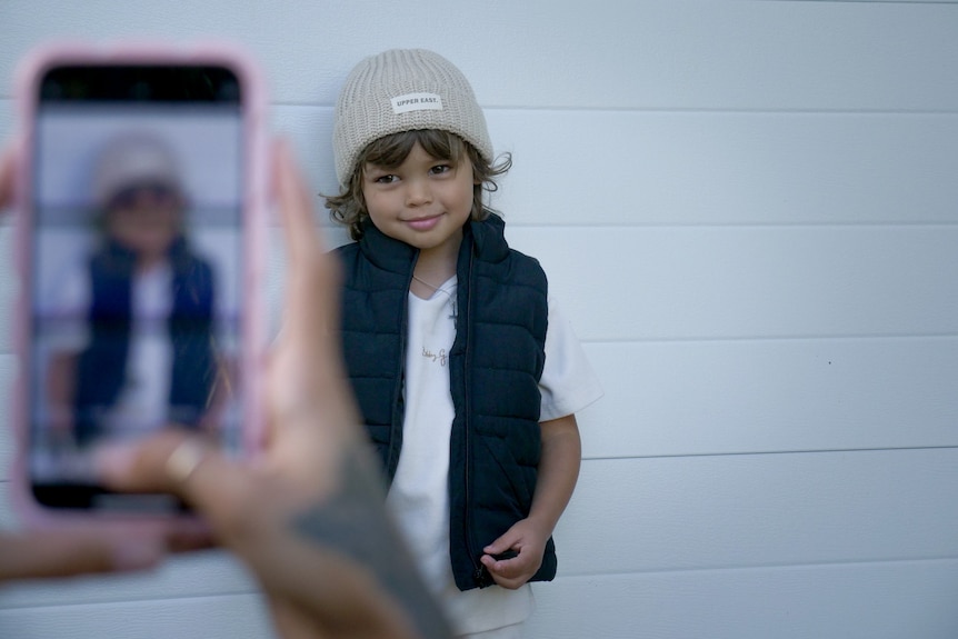 A young boy standing next to a wall and smiling while someone takes a photo on a pink iPhone.
