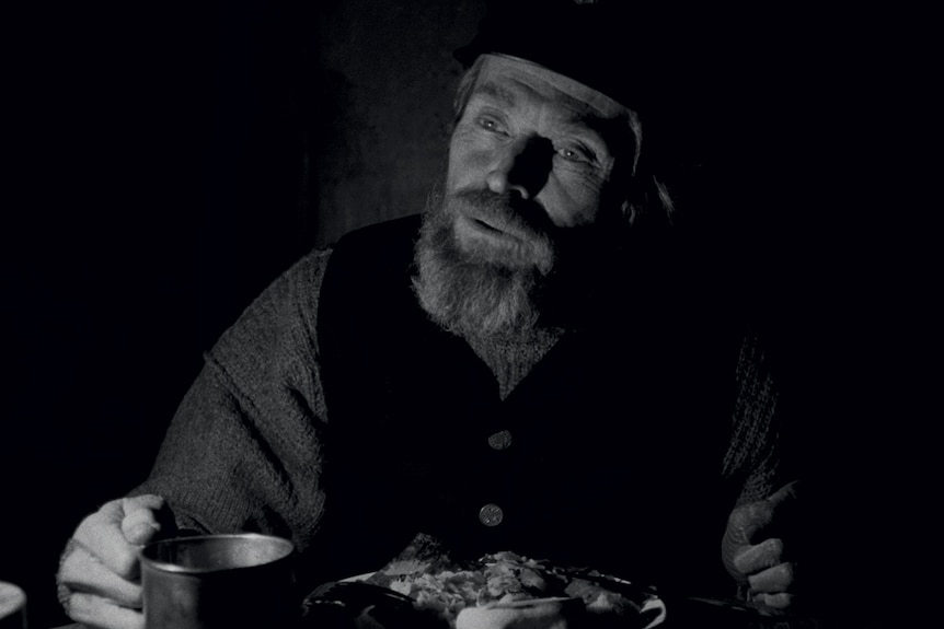 Black & white image from the movie the Lighthouse featuring Willem Dafoe looking tired and haggard and eating a meal