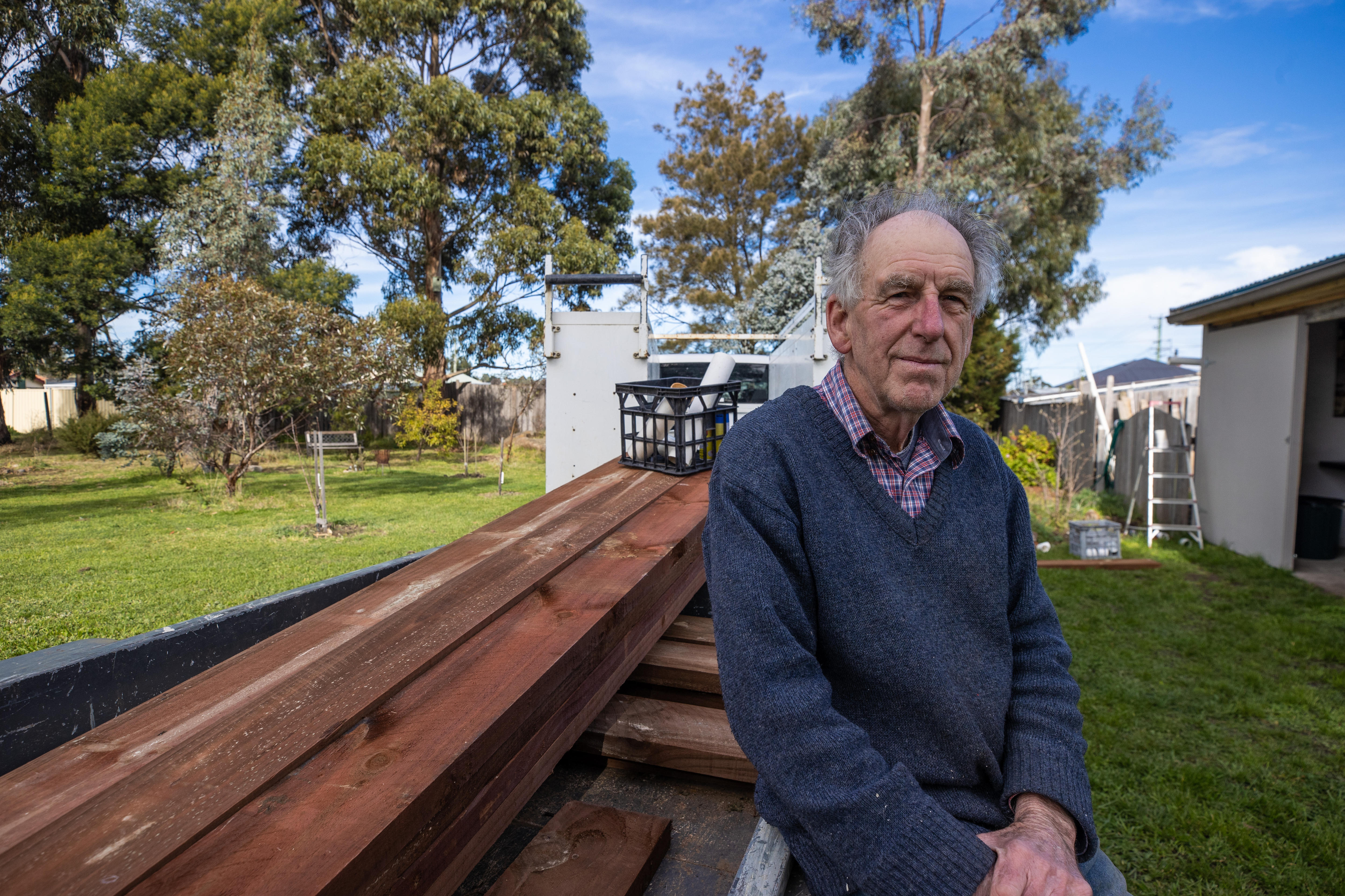 An older man smiles at the camera while leaning against a pile of timber.