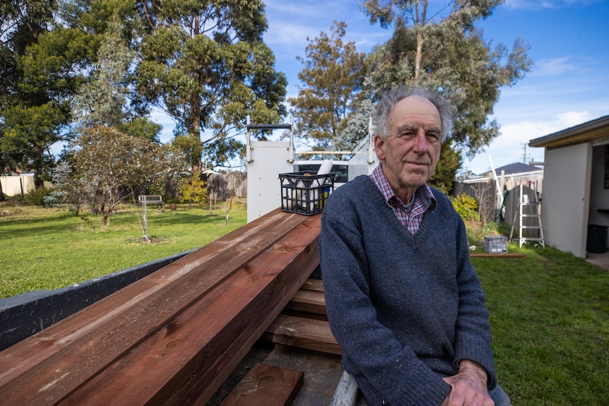 An older man smiles at the camera while leaning against a pile of timber.