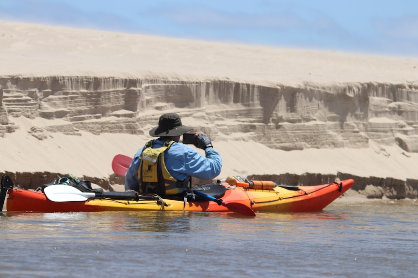 A mid-shot from behind of Brenton, a fair-skinned man, taking a photo from his kayak next to a sand mound. 