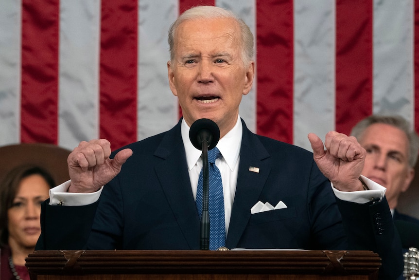 Joe Biden gestures with his thumbs at a lectern in front of a large American flag.