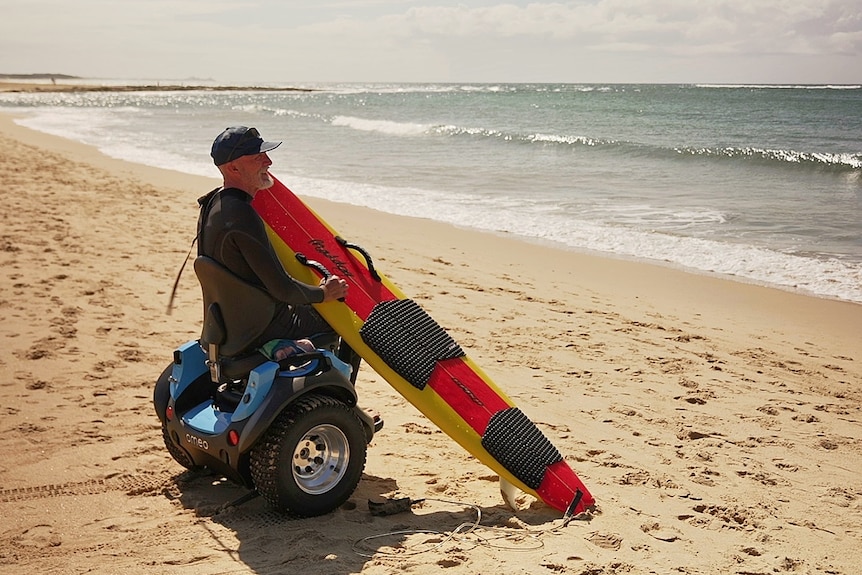 Colin in his wheelchair on the beach with his surfboard