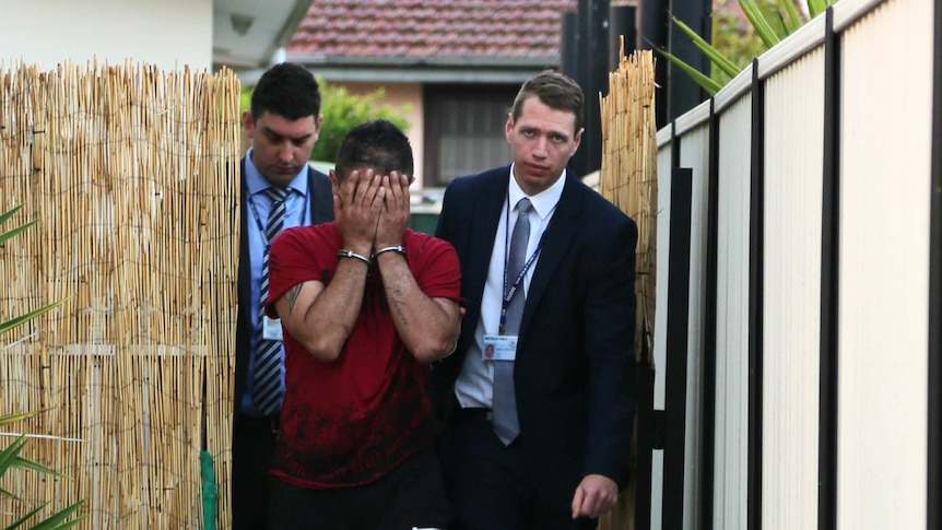 Detectives lead a man in handcuffs through a bamboo screen fence.